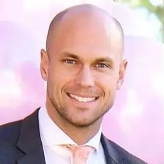 Smiling bald man in a dark suit jacket, white shirt, and light pink tie in front of a pink balloon backdrop.