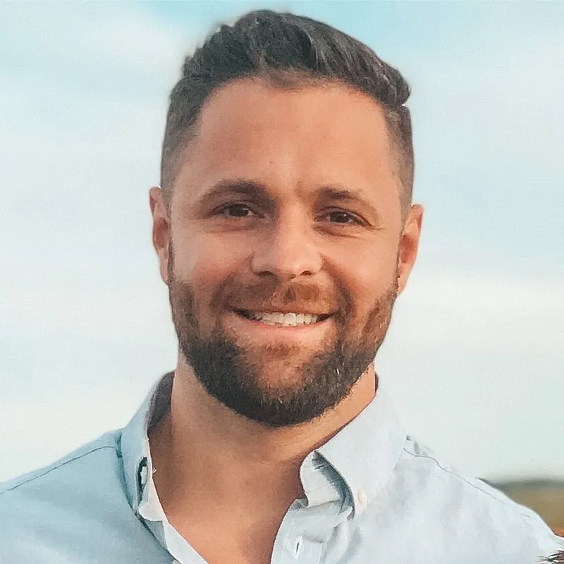 Smiling man with short dark hair and beard wearing a light blue button-up shirt against a blue sky background.